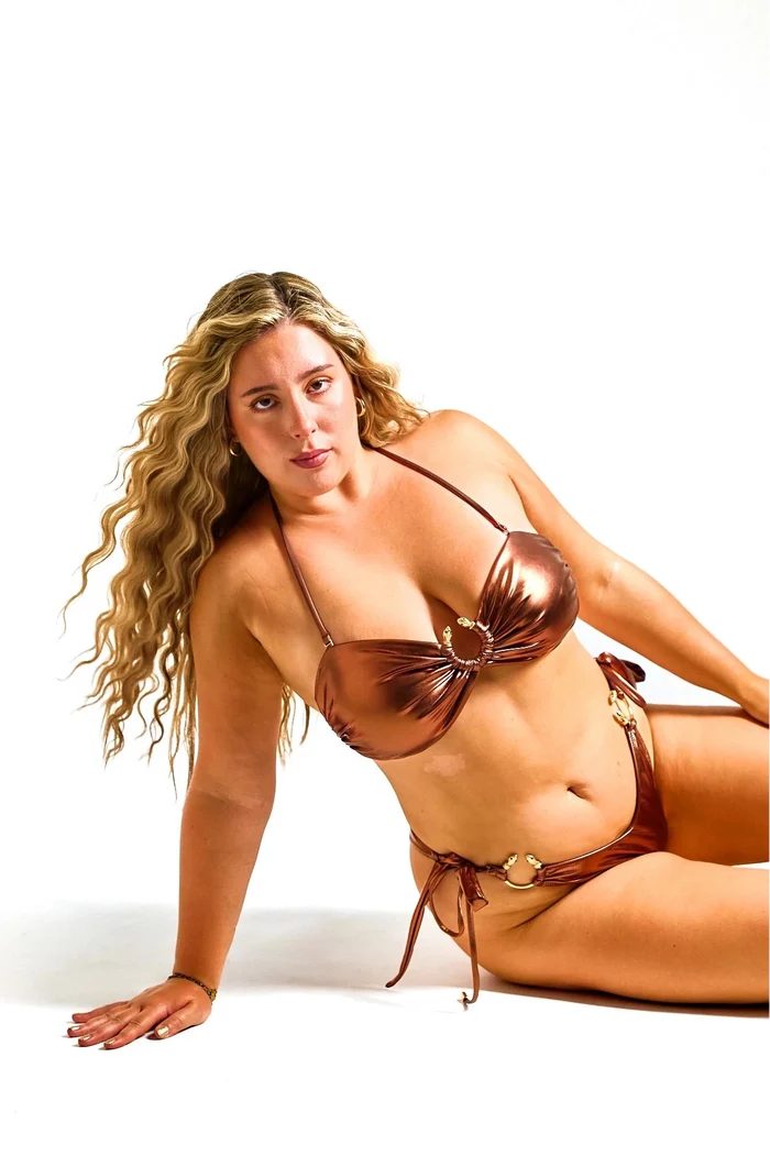 A woman with long curly blonde hair poses on a white studio floor wearing a metallic bronze bikini with gold-toned snake details.